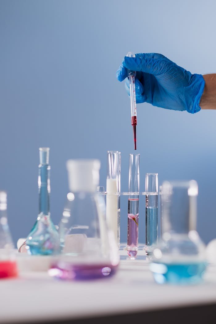 A scientist's hand with a glove holding a dropper above test tubes with colorful liquids in a lab setting.